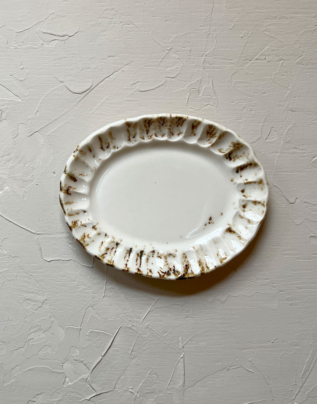 The Oval Side Plate from the new DBO HOME Nonna collection is a glossy organic off-white ceramic plate with an oval shape and a slightly raised fluted, ruffled edge, adorned with painterly drips and splashes of bronze glaze around the iconic rim. It measures 8.5 inches by 6.5" and sits alone on a plastered, sand-colored surface.