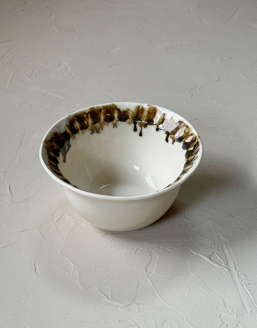 The Small Bowl from the new DBO HOME Nonna collection is a glossy organic off-white ceramic bowl with a deep belly and a fluted, ruffled edge, adorned with painterly drips and splashes of bronze glaze around the iconic rim. It measures 6 inches in diameter and 3 inches in height and sits alone on a plastered, sand-colored surface.